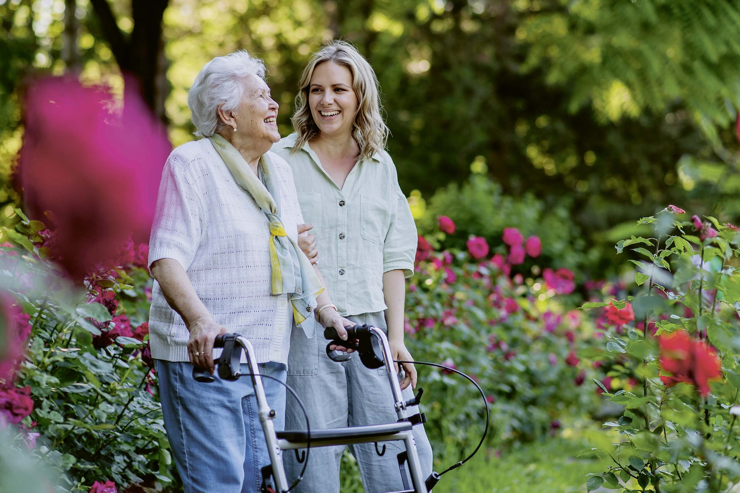 Eine junge Frau läuft im Garten neben einer älteren Frau am Rollator.