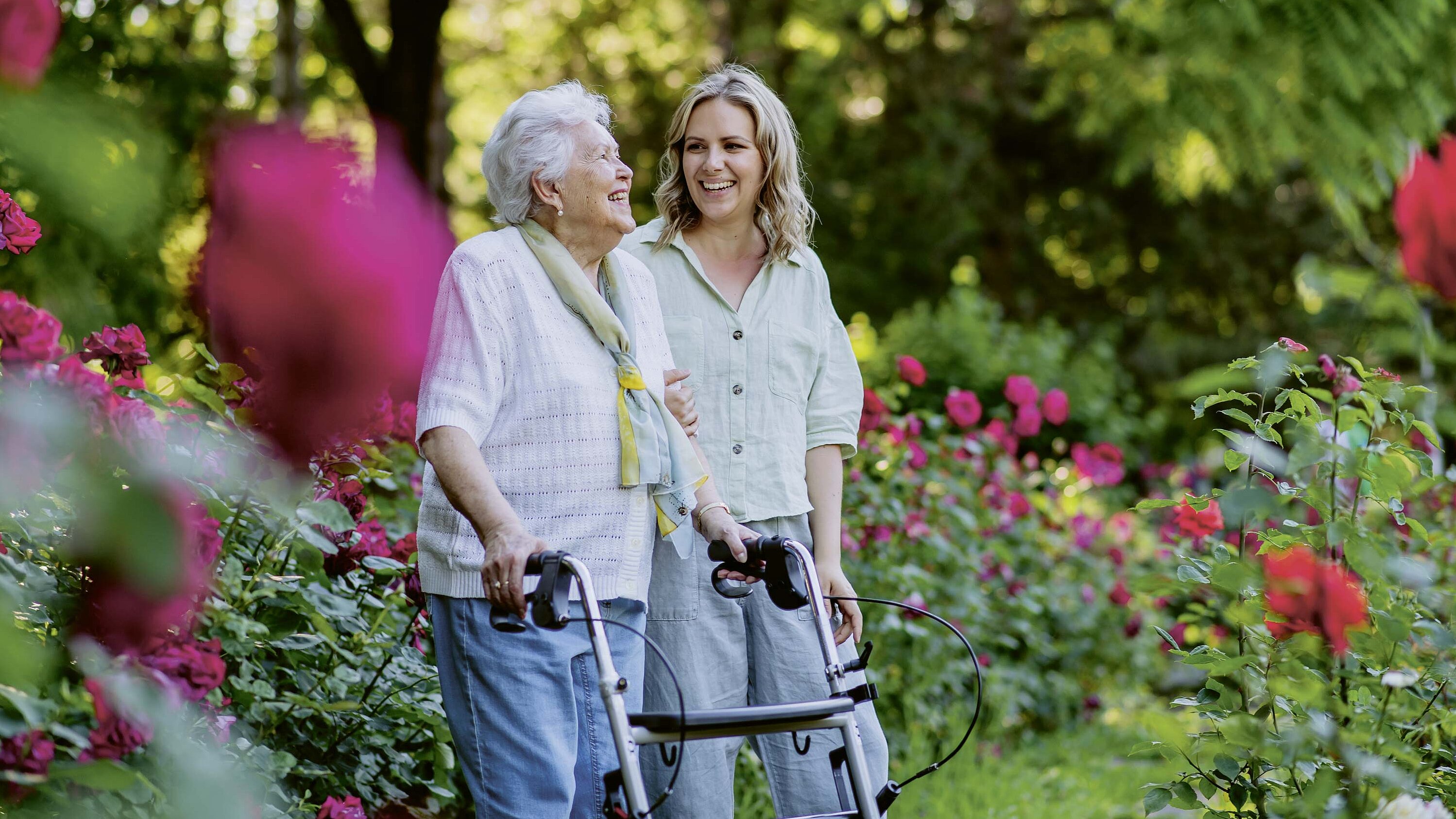 Eine junge Frau läuft im Garten neben einer älteren Frau am Rollator.