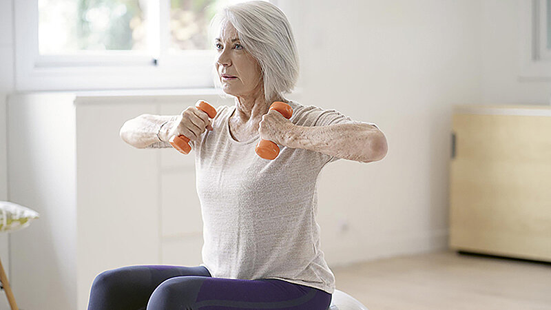 Eine ältere Frau sitzt in Trainingskleidung auf einen Gymnastikball und trainiert ihre Arme mit ein- bis zwei Kilo Hanteln.