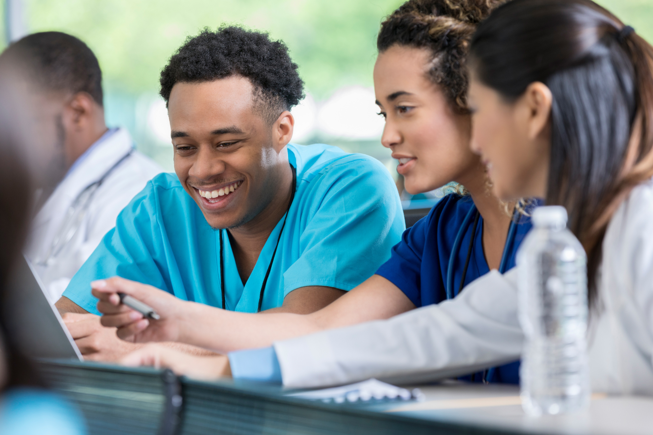 Ein junger Pfleger und zwei junge Pflegerinnen sitzen an einem Tisch und schauen gemeinsam in einen Laptop währenddem sie interessiert lachen.