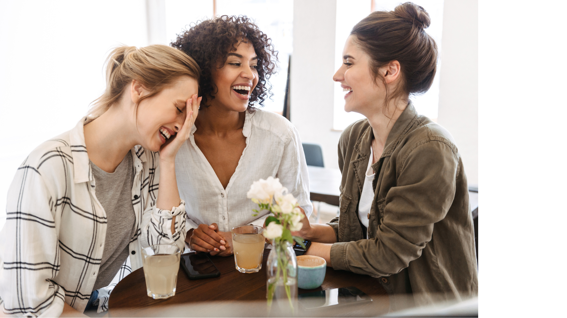 Drei Frauen sitzen an einem Tisch und trinken Kaffee. Sie lachen ausgelassen.