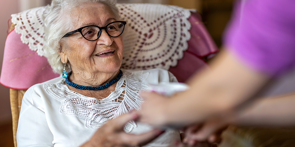 Eine ältere Frau in einem Sessel hält lächelnd die Hände einer Person von der nur die Arme zu sehen sind.