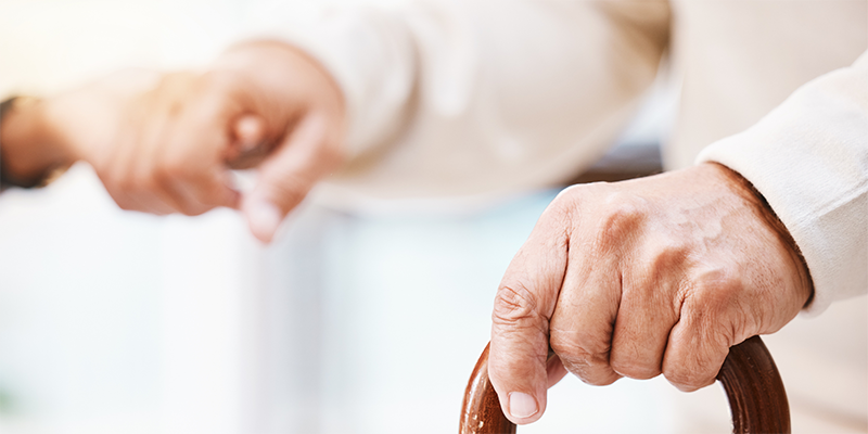 Die Hände eines älteren Mannes stützen sich links auf einem Gehstock ab und werden rechts von einer weiteren Hand gehalten