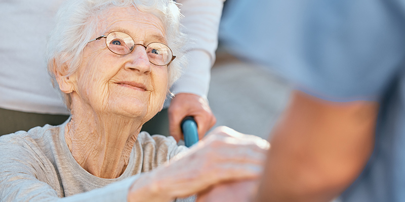 Eine ältere Frau im Rollstuhl lächelt zufrieden eine Person an, von der im Bild nur die Arme zu sehen sind, und hält deren Hände.