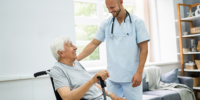Ein junger Pfleger mit Glatze steht neben einem älteren Mann im Rollstuhl und greift ihm mit der Hand an die Schulter währenddem die beiden sich lachend anschauen.