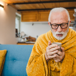 Älterer Mann sitzt in einer Decke mit einem Becher Tee auf dem Sofa