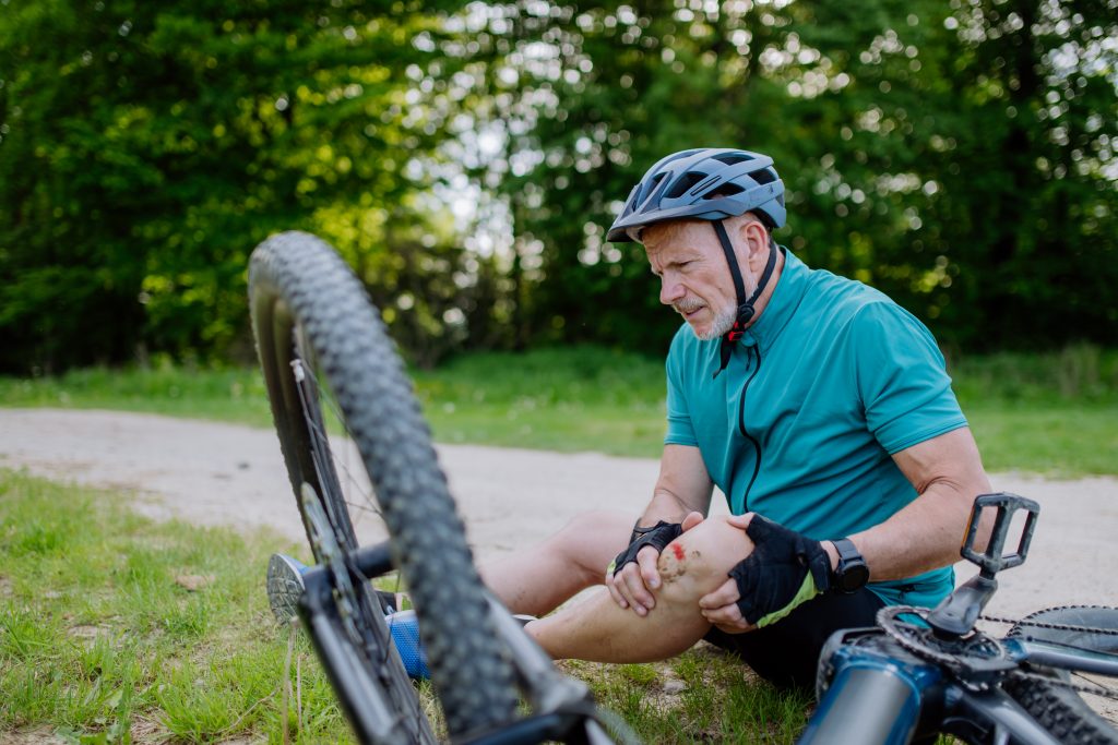 Ein Senior in Sportkleidung ist von seinem Fahrrad gefallen und sitzt jetzt auf dem Boden. Sein Knie ist verletzt.