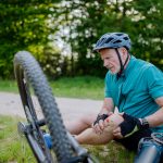 Ein Senior in Sportkleidung ist von seinem Fahrrad gefallen und sitzt jetzt auf dem Boden. Sein Knie ist verletzt.