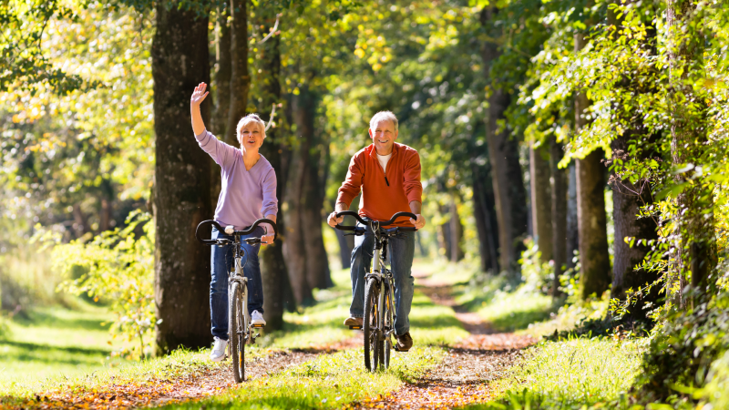 Radfahren für Senioren Zwei Senioren fahren durch einen Wald auf dem Fahrrad. Die Sonne scheint und sie wirken fröhlich.