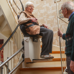 2 ältere Meschen, ein fährt mit dem Treppenlift, die andere schaut ihr dabei zu.