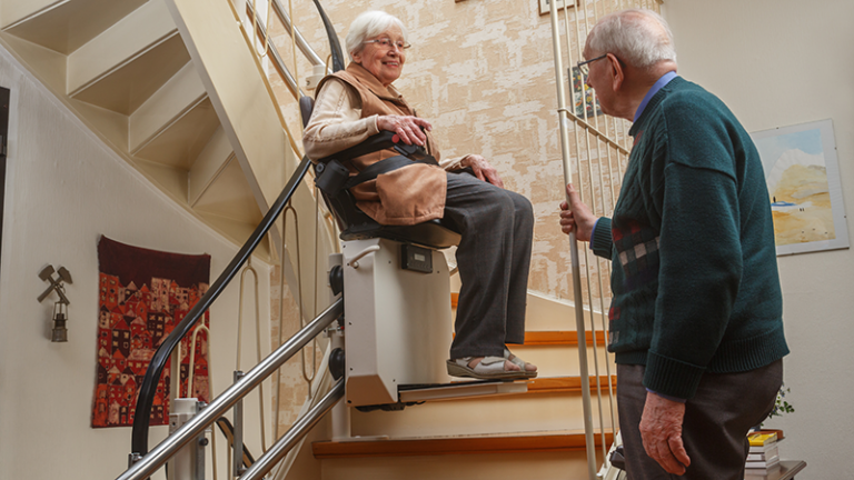 2 ältere Meschen, ein fährt mit dem Treppenlift, die andere schaut ihr dabei zu.