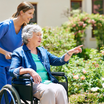 Pflegerin schiebt Bewohnerin auf dem Rollstuhl durch den Garten
