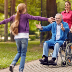 Tochter rennt Opa in die Arme. Er sitzt auf einem Rollstuhl und wird von einer Pflegerin gestossen