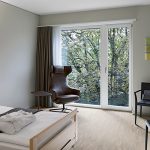 Stylisches Zimmer im Pflegezentrum mit Bett, Ohrsessel, Tisch und Aussicht durch ein grosses Fenster auf einen Baum