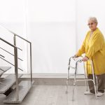 Ältere Frau steht mit Gehilfe unter einer Treppe