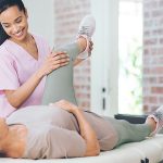 Physiotherapeutin hebt Bein von älterer Frau auf einer Liege