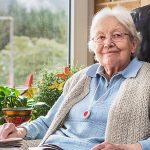 Ältere Frau sitzt in einem Sessel am Fenster und liesst Zeitung